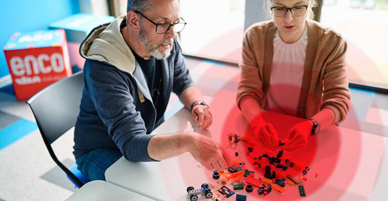 weißer Tisch mit Bausteinen in verschiedenen Varianten und Farben und zwei Personen, die die Variantensteine in der Hand halten., die die Variantenvielfalt widerspiegeln soll Zudem ist ein Schmerzsymbol über den Bausteinen auf dem Tisch zu erkennen.