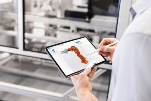 Ein Mann im weißen Hemd stellt auf einem Tablet einen orangefarbenen Roboterarm mittels 3D-Konfiguration zusammen. Im Hintergrund ist schemenhaft eine Fabrikhalle zu sehen.