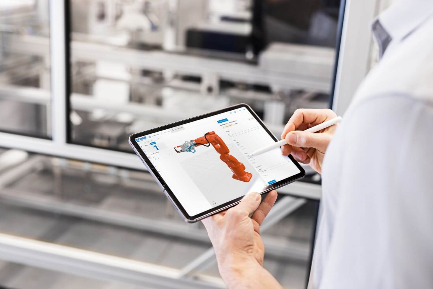 Ein Mann im weißen Hemd stellt auf einem Tablet einen orangefarbenen Roboterarm mittels 3D-Konfiguration zusammen. Im Hintergrund ist schemenhaft eine Fabrikhalle zu sehen.